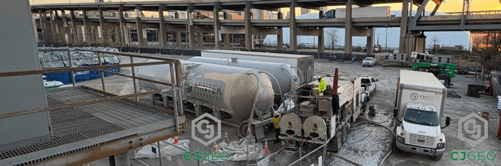 A CJGeo cellular grout batch plant, support truck and two cement pigs sit in a gravel yard next to a double layer bridge.