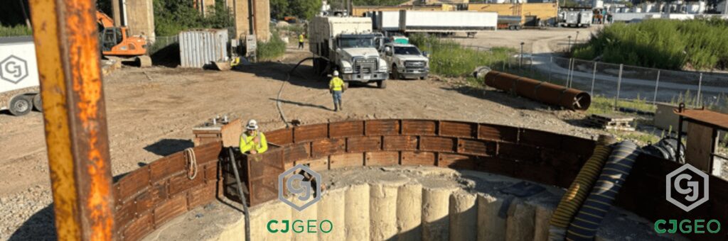 A wide view of a CJGeo construction site, focusing on the top of a large diameter augered secant pile shaft. A worker in a hard hat stands near the edge of the shaft, which is ringed by a curved steel liner plate structure. In the background, an excavator, grout plant, and other site activity are visible.