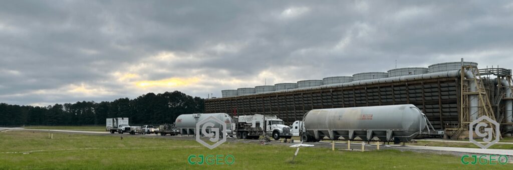 A long shot of CJGeo's cellular grout trucks and bulk cement tankers lined up on an access road to perform a pipe abandonment job, with the massive cooling towers of a power plant in the background under an overcast sky.
