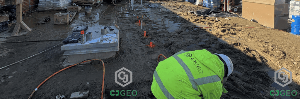 A CJGeo worker leaned over on a muddy jobsite with manchette tubes sticking out of the ground for permeation grouting.